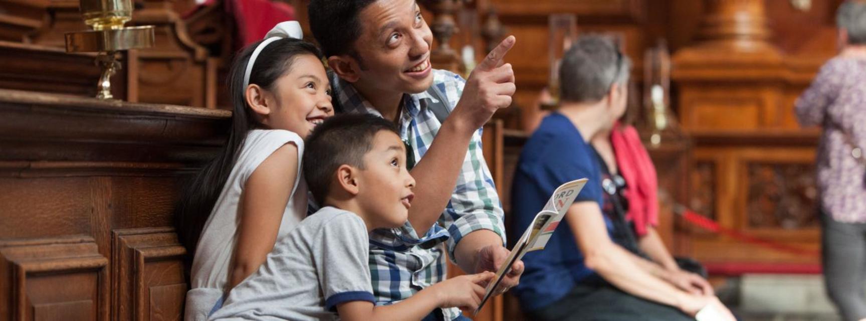 Photo of a family sitting inside the Sheldonian theatre, holding the Oxford Open Doors brochure and pointing, looking and smiling at the ceiling 