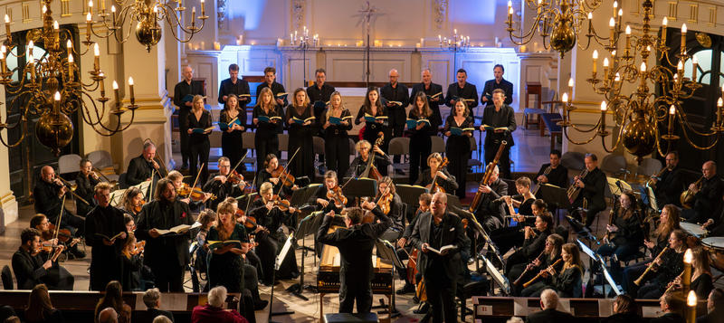 La Nuova Musica performing company mid performance at an indoor venue, lit by chandeliers and blue uplighters behind them