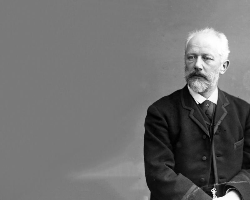 Tchaikovsky sits with his hands in his lap, waring a velvet jacket and cravat. He has a beard and moustache and looks thoughtfully into the distance.