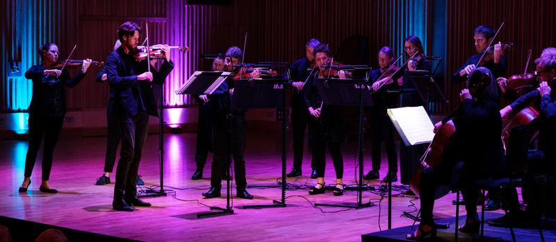 Britten Sinfonia strings stood performing on a stage lit with purple and blue uplighters