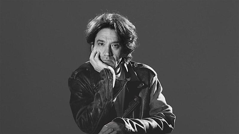 Curtis Yarvin sits against a black background looking straight ahead and rests his chin on his right hand.