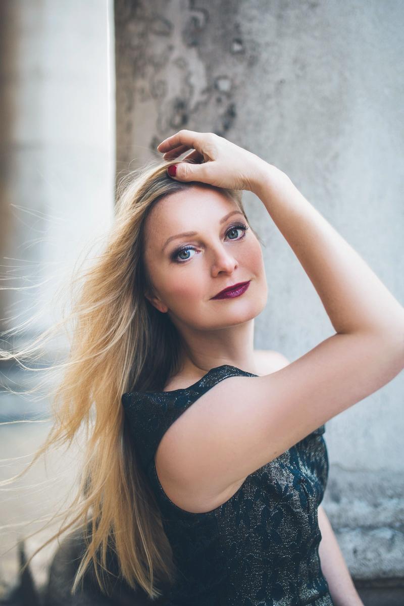 A blonde haired woman stands with her back to the camera, looking over her shoulder and smiling with her arms crossed above her head.