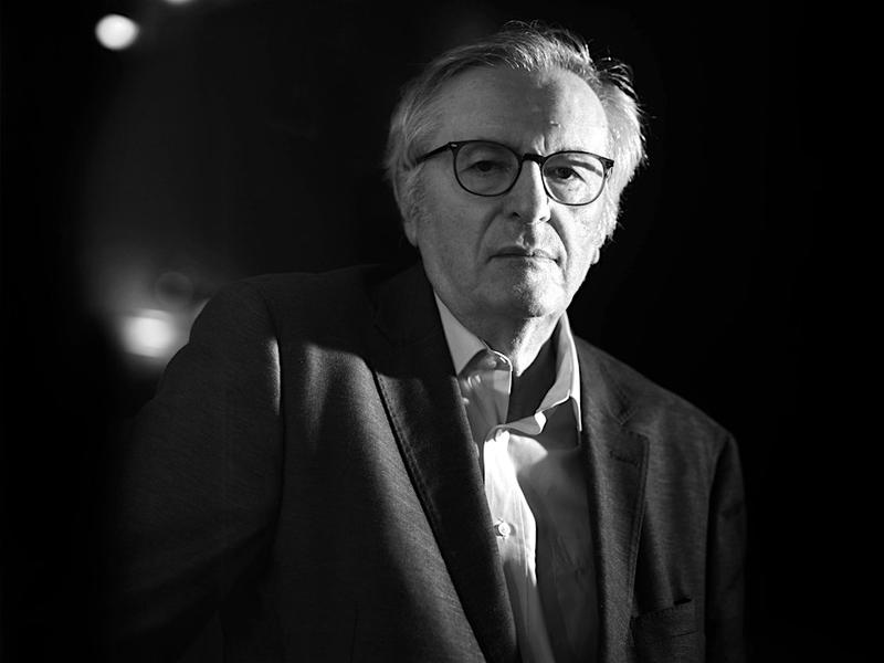 John Gray stands in a jacket and shirt, wearing glasses and looking straight ahead. 