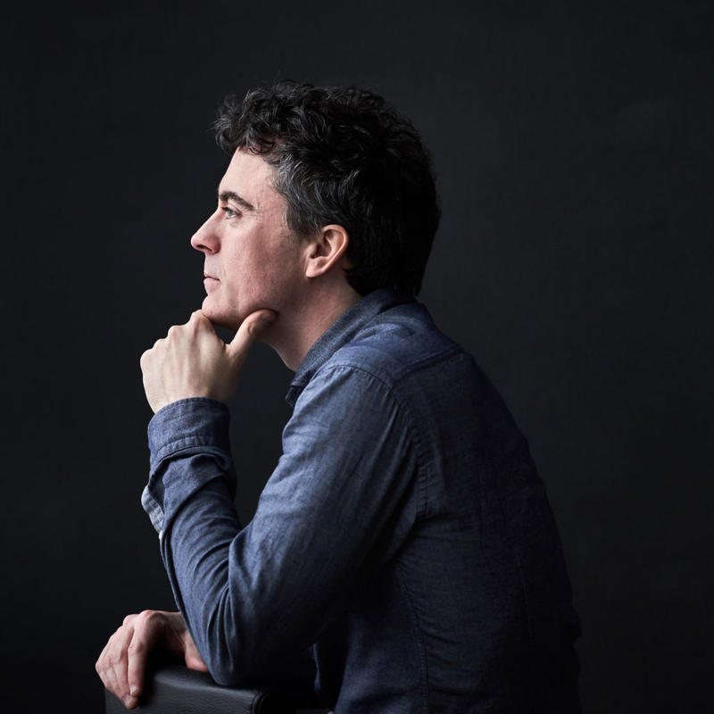 A side profile of a man in a long sleeved blue shirt against a black background, looking pensively to the left