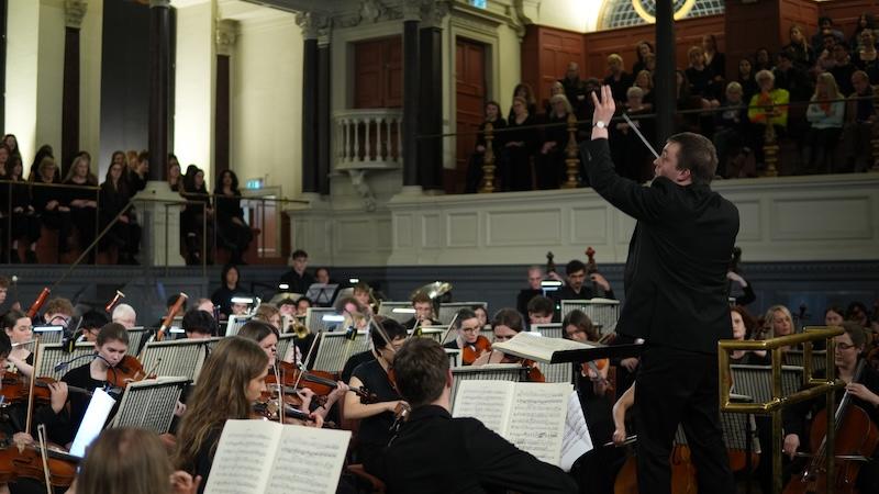 Conductor Joe Davies conducting the Oxford Millennium Orchestra in an evening-lit Sheldonian Theatre