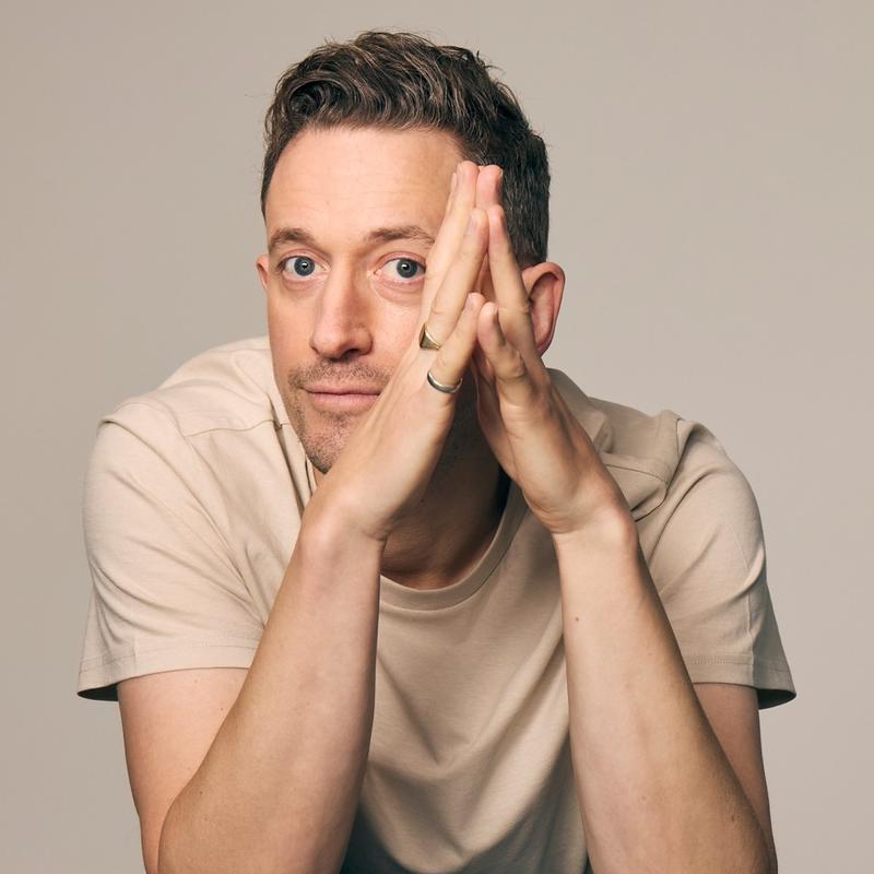 John Robins, who has short dark hair, sits with his fingertips touching in front of his face and elbows resting on a grey table in front of him.
