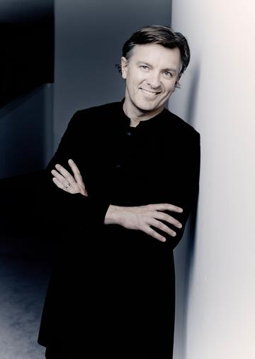 A man stands leaning against a black wall, smiling at the camera with his arms folded.